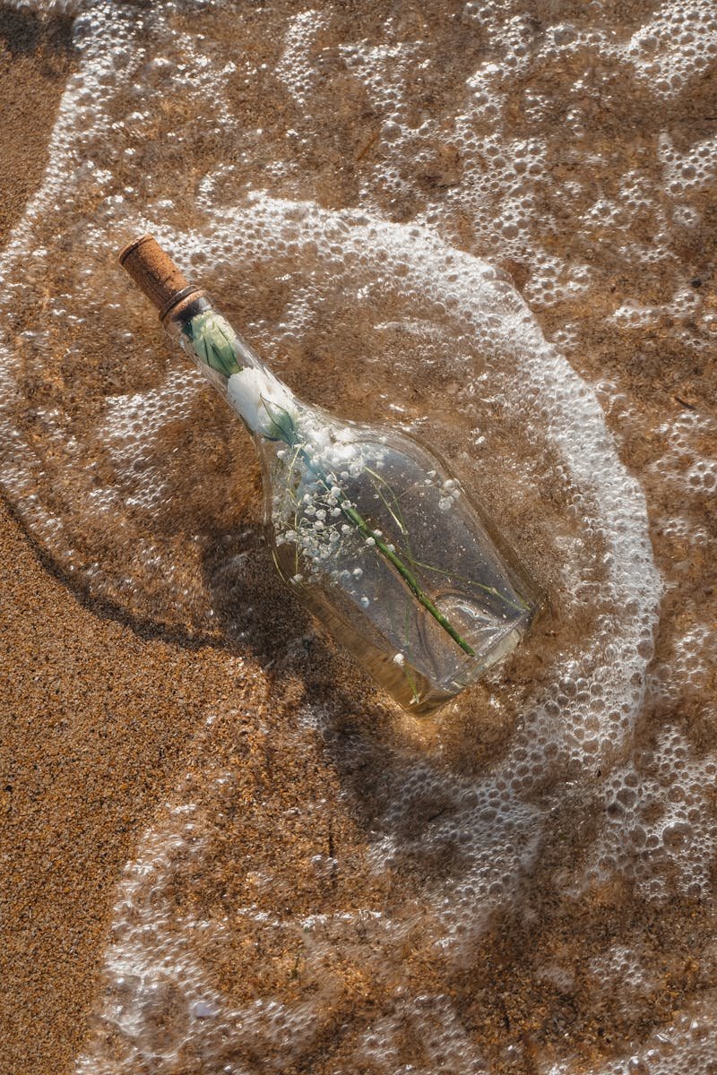 Glass bottle with flowers surrounded by gentle waves on a sandy beach.